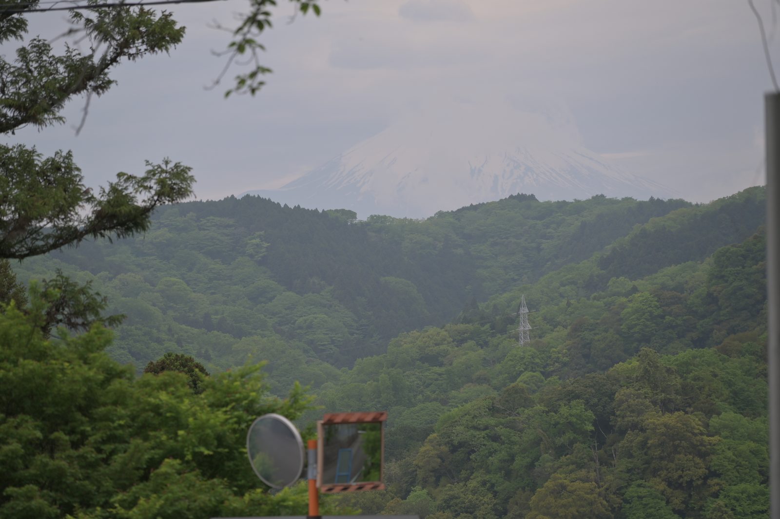 下山途中にふいに現れた富士山。曇り空の中、緑の山並みの向こうに雪をかぶった富士山がうっすら見える。