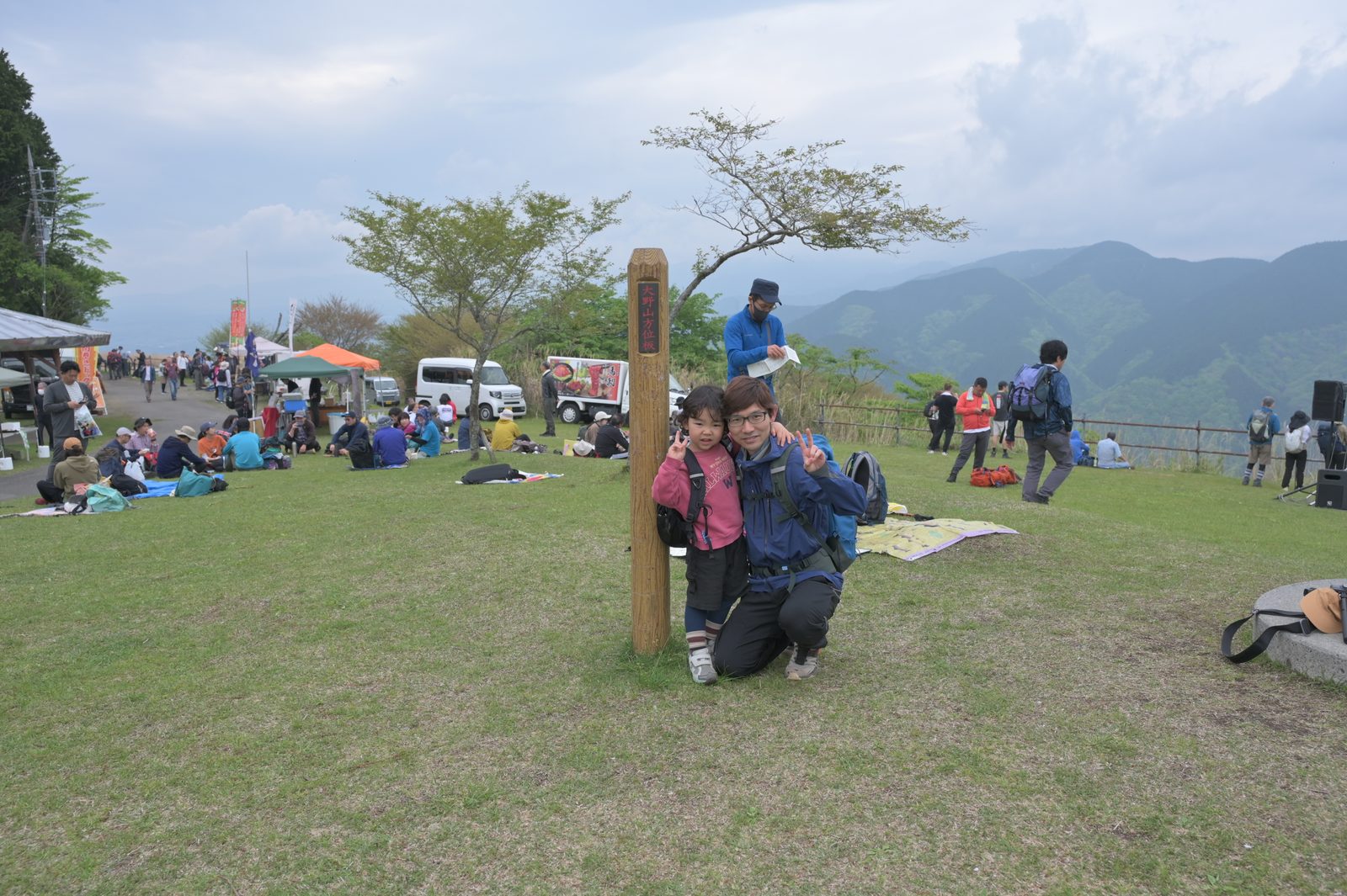 山頂標識の前でポーズをとる親子。背後には山開きイベントのテントと多くの登山者、丹沢の山並みが広がる。