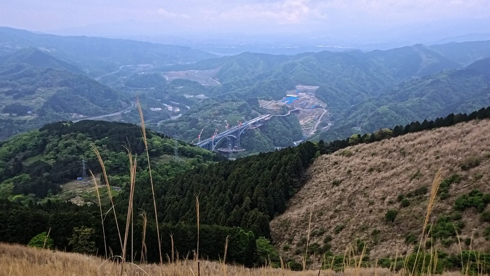 大野山の稜線から見下ろす眺望。山間に工事中の大きなアーチ橋と緑の山並みが広がる。