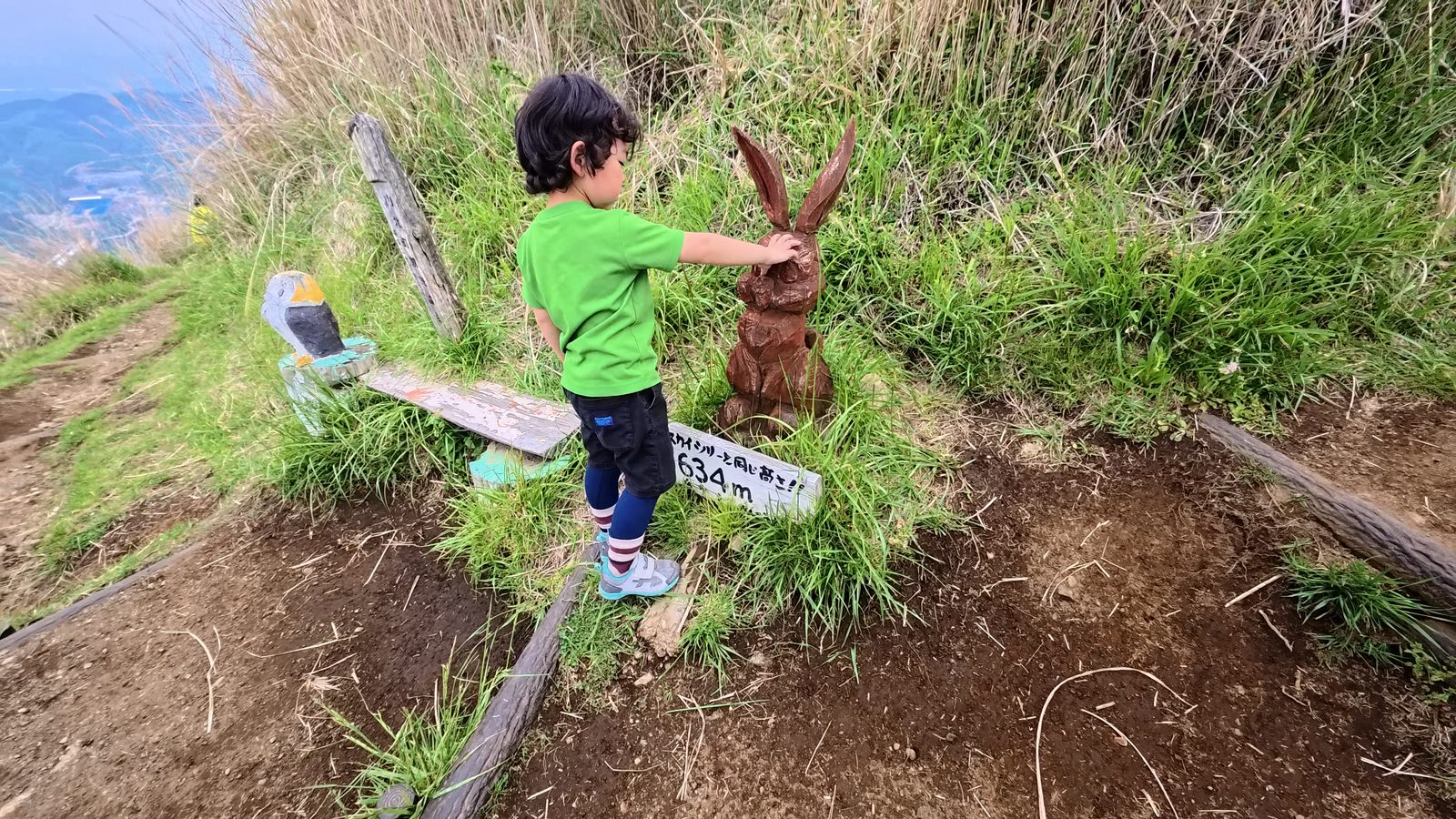 「スカイツリーと同じ高さ 634m」の看板の前で、子供がウサギのチェンソーアートに手を伸ばしている。